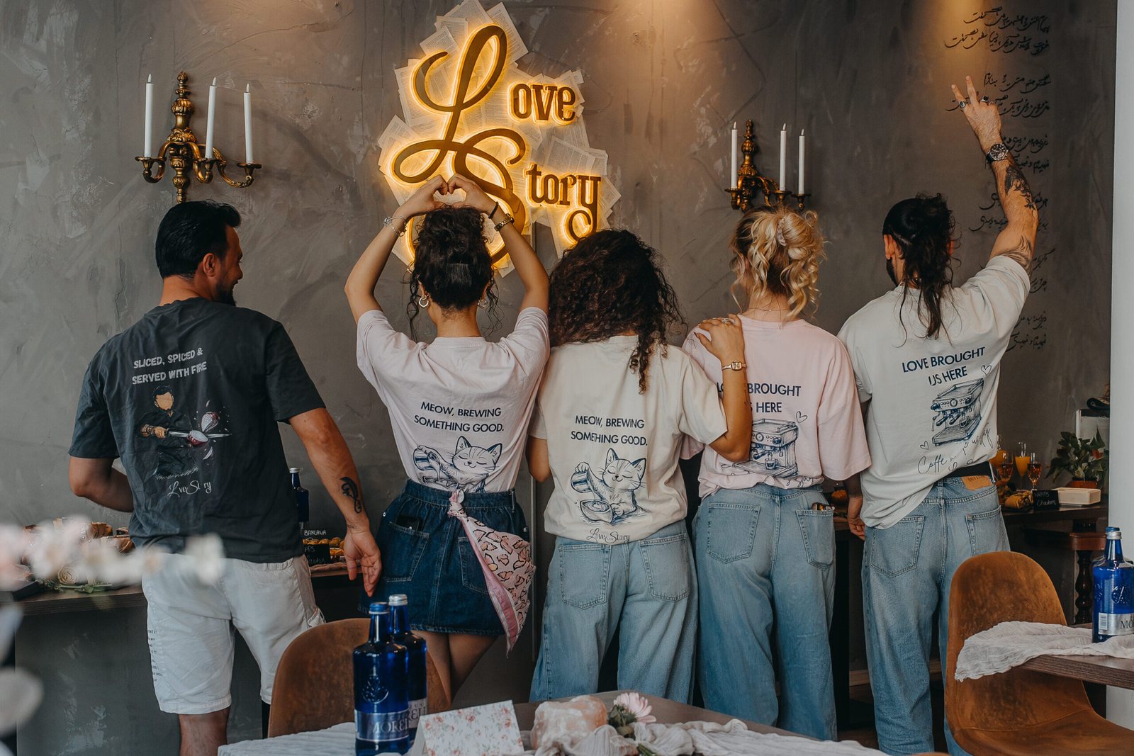 Das Team des Café Love Story steht mit erhobenen Armen vor dem gelben Neon-Schild an der Betonwand